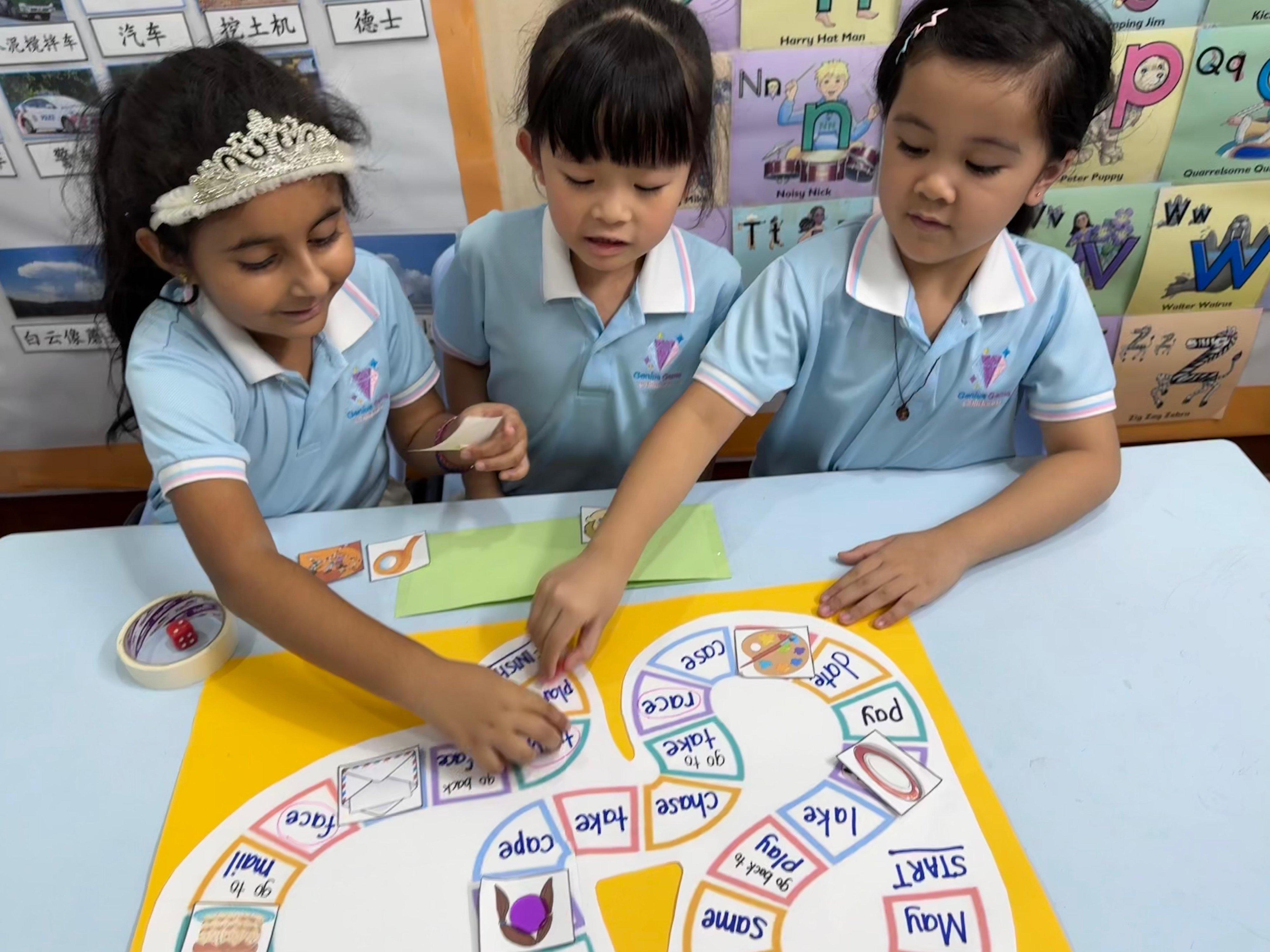 Three children playing a hand-made word board game together at a classroom table at Genius Gems Childcare Centre