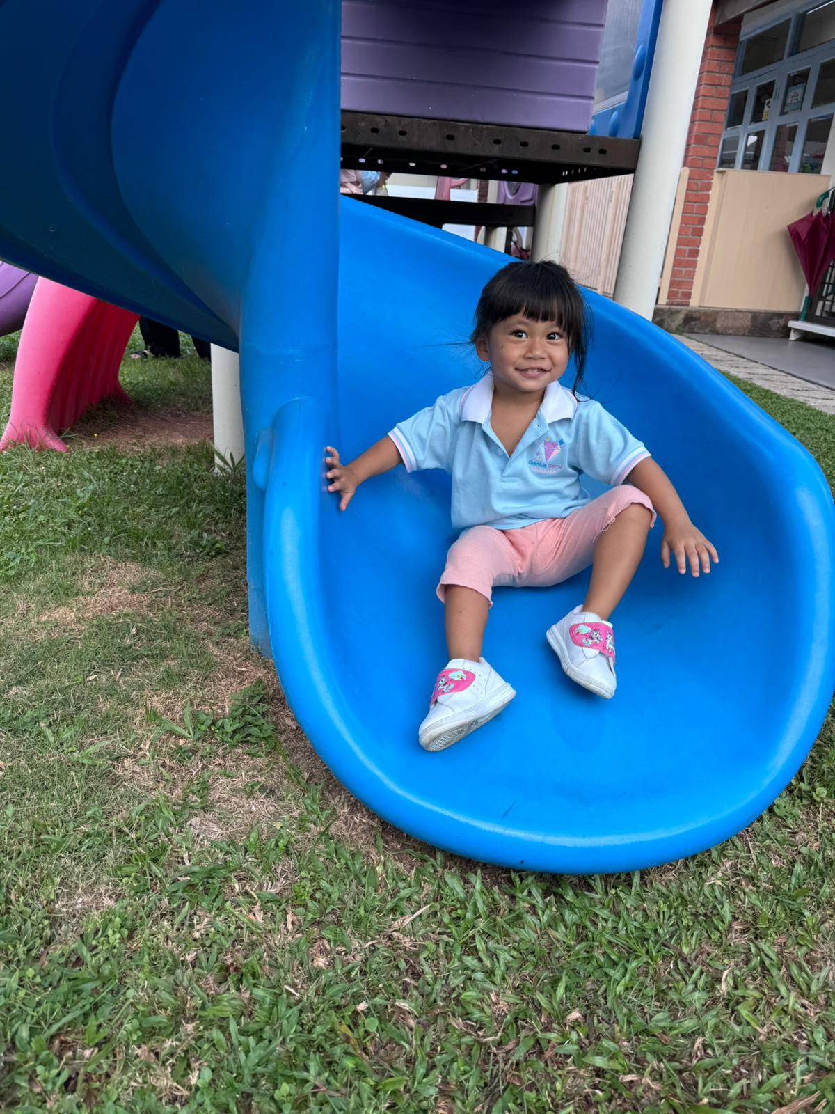 A smiling Genius Gems child sliding down the big blue slide on the large outdoor playground